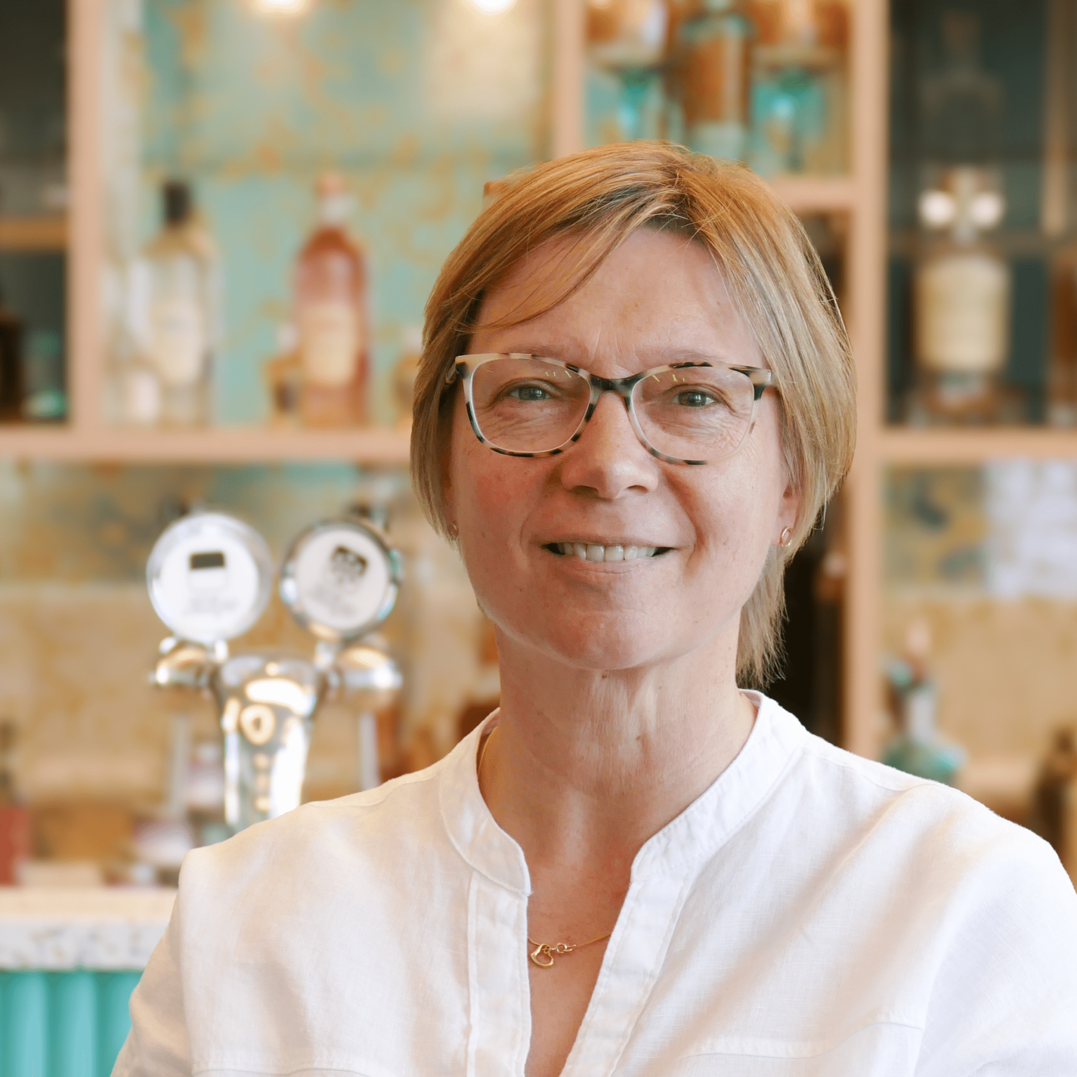 A woman with short, blonde hair and glasses, wearing a white shirt, standing in front of shelves filled with bottles.