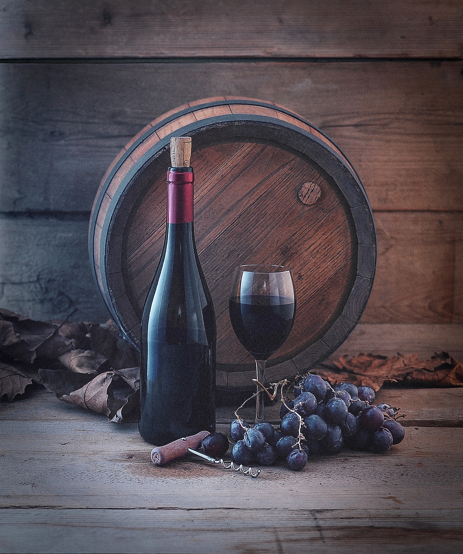 Wine bottle, glass, and grapes in front of a wooden barrel on a rustic table.