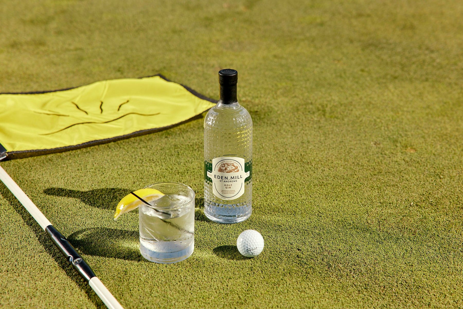 An Eden Mill Golf Gin Bottle on a golf green with a yellow flag and a golf ball.